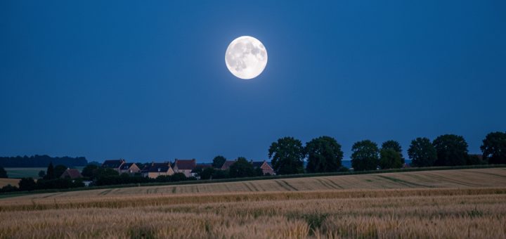 Une pleine lune lumineuse se lève au-dessus d’un paysage français estival sous un ciel bleu profond au crépuscule.